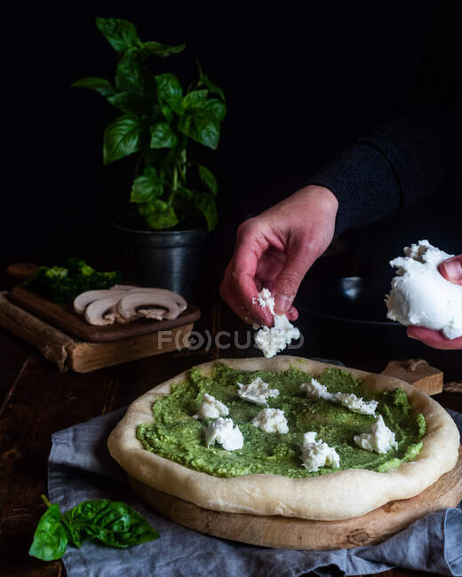 Crop chef anônimo adicionando saboroso queijo Mozzarella na pizza com molho pesto e preparar o almoço na mesa escura — Fotografia de Stock