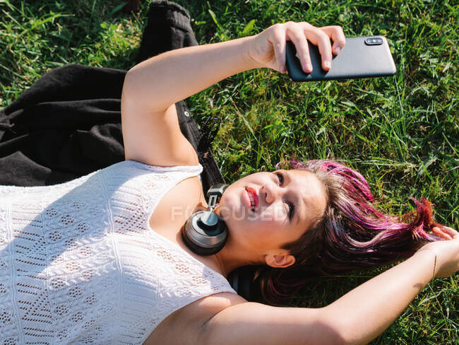 Von oben eine unbeschwerte, pummelige Teenagerin mit Kopfhörern, die sich im Gras entspannt und im sonnigen Park ein Selbstporträt auf dem Handy macht — Stockfoto