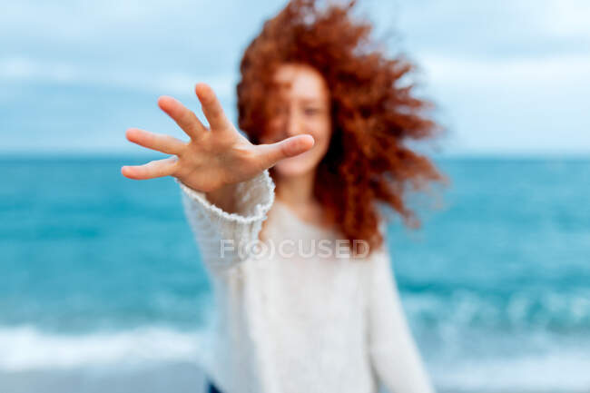 Приятная женщина с длинной рыжей кудрявой прической, протягивающей руку к камере, стоя напротив моря. — стоковое фото
