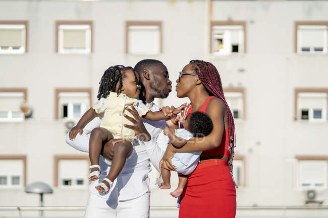Весела афроамериканська жінка з плечима й немовлятком на руках стоїть і цілує чоловіка з позитивною донькою на руках на вулиці проти житлового будинку в сонячний день. — стокове фото