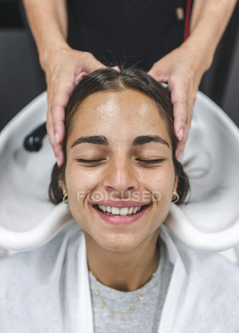 Cultiver maître méconnaissable laver les cheveux foncés du client féminin avec les yeux fermés dans le salon de beauté — Photo de stock