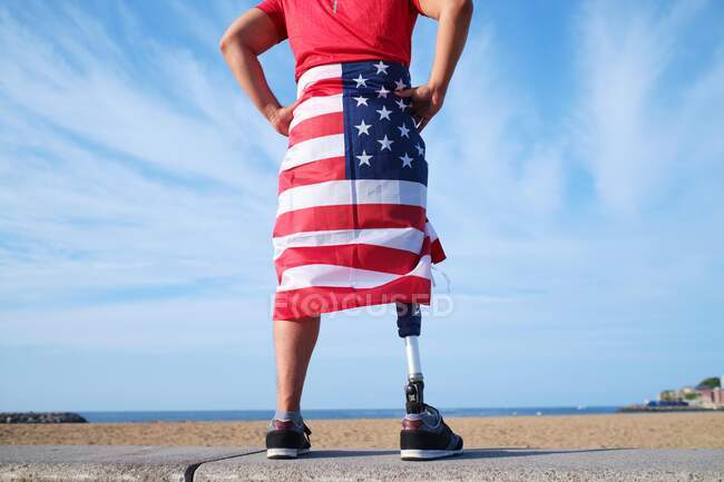 Vista posteriore di sportivo irriconoscibile con protesi alle gambe in piedi con bandiera americana avvolta intorno al fianco sul bordo di cemento sopra un cielo con nuvole — Foto stock