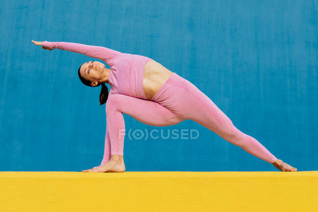 Slim fêmea em sportswear rosa praticando ioga em Utthita Parshvakonasana no fundo azul brilhante e amarelo — Fotografia de Stock