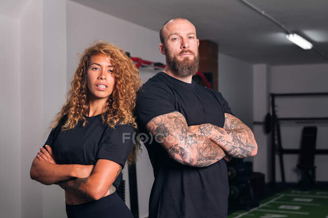 Confident muscular sportive instructors standing with crossed arms in gym and looking at camera in daytime — Stock Photo