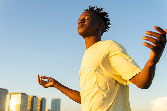 D'en bas de calme Afro-Américain mâle en vêtements décontractés debout avec les bras levés et les yeux fermés sur la rue en été — Photo de stock