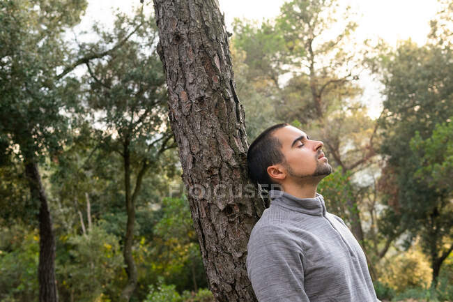 Side view of peaceful young ethnic male hiker in casual clothes and rucksack leaning on tree trunk with closed eyes and enjoying fresh air of green forest in mountainous valley — Stock Photo