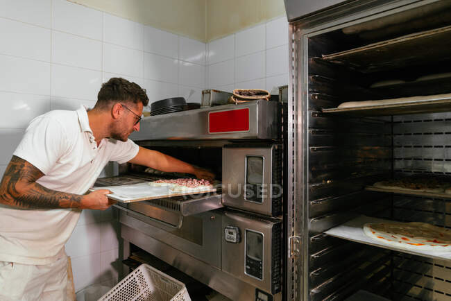 Side view of concentrated man chef in casual clothes and eyeglasses standing and putting baking pan with raw pizza in oven in kitchen — Stock Photo
