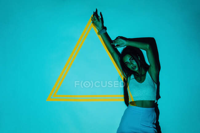 Модна етнічна жінка тисячоліття з довгим волоссям і піднятою рукою дивиться на камеру проти жовтого трикутника з проектора світла — стокове фото