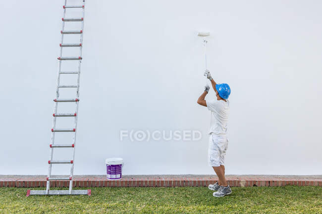 Joven pintor irreconocible pintando la fachada de un apartamento con un rodillo con la escalera a un lado - foto de stock