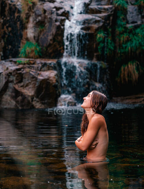 Vista lateral de sonriente mujer desnuda viajando cubriendo pecho y de pie en aguas tranquilas de estanque cerca de cascada en el parque natural Fervenza De Casarinos en España - foto de stock