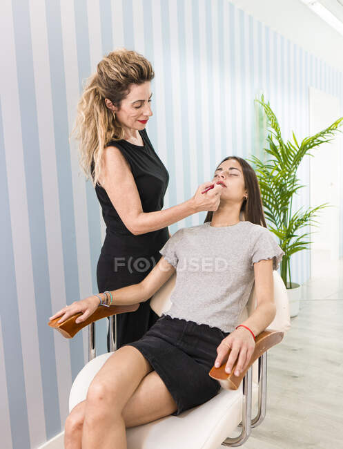 Side view of crop visagiste applying makeup with lipstick on the lips of young female — Stock Photo