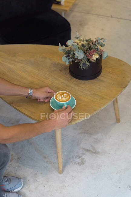 Crop anonymous person putting mug of hot coffee with latte art on wooden table with decorative flowers in coffee house — Stock Photo