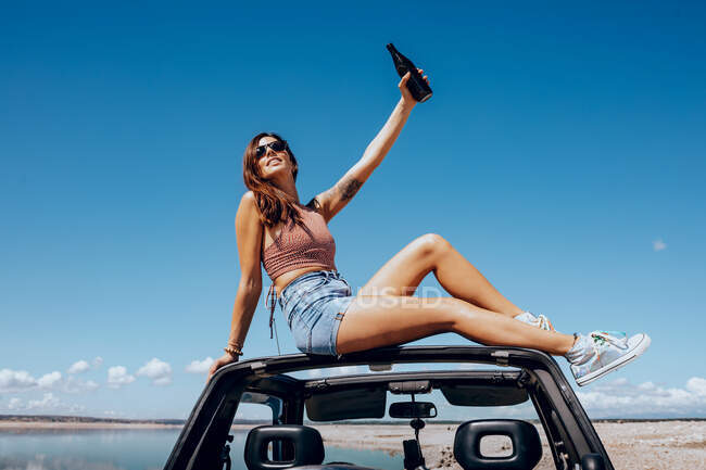 Cuerpo completo de feliz joven hembra en traje de verano y gafas de sol levantando la mano de la cerveza mientras está sentado en el techo del coche safari en la orilla del río - foto de stock
