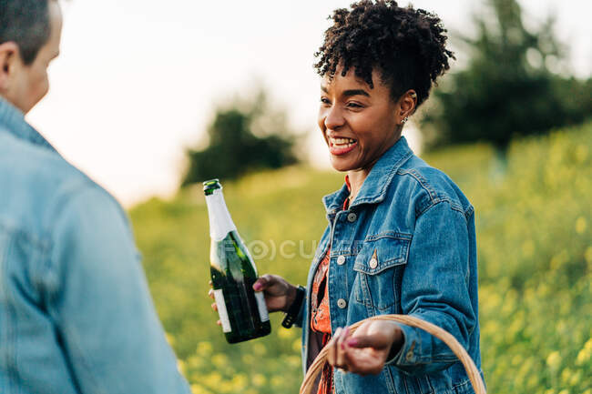 Cheerful young black female with curly hair in stylish dress and denim jacket smiling happily and looking at crop unrecognizable boyfriend while standing in nature with basket and champagne bottle — Stock Photo