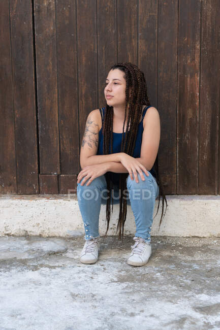 Cuerpo completo de mujer hispana tranquila en jeans con pelo largo trenzado mirando a la distancia mientras está sentada cerca de la pared de madera en la calle - foto de stock