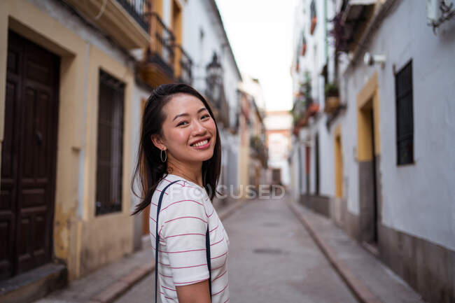 Вид сбоку улыбающейся азиатской туристки, стоящей в узком проходе между домами, глядя на камеру в городе — стоковое фото