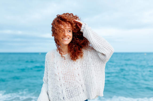 Délicieuse femelle avec de longs cheveux bouclés au gingembre portant un pull tricoté debout regardant loin contre la mer bleue — Photo de stock