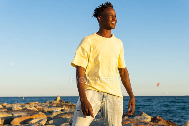 Happy African American male in casual clothes standing on rocky seashore while smiling and looking away in summer evening — Stock Photo