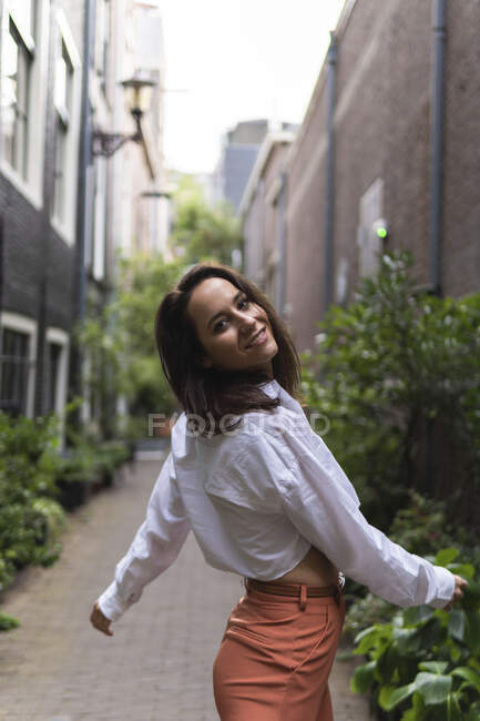 Attractive female in casual clothes walking on narrow street near buildings and plants while looking at camera — Stock Photo