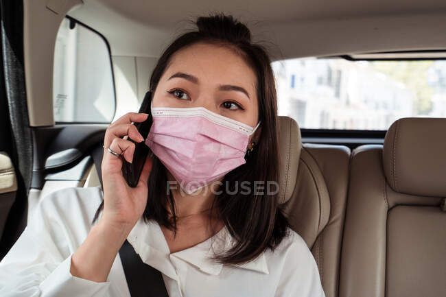 Feche o passageiro feminino étnico em máscara protetora sentado com cinto de segurança e ter chamada telefônica em táxi — Fotografia de Stock