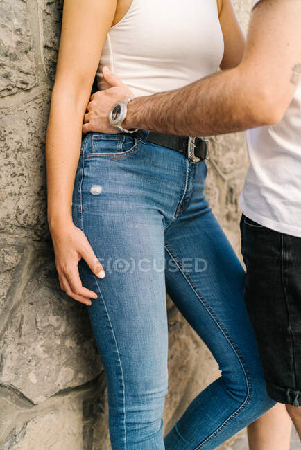 Side view of anonymous young ethnic man in casual clothes embracing girlfriend leaning on aged stone building in city park on sunny day — Stock Photo