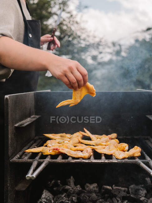 Crop anonymous chef putting raw chicken wing on hot metal grill grate with smoke while cooking in countryside during picnic — Stock Photo