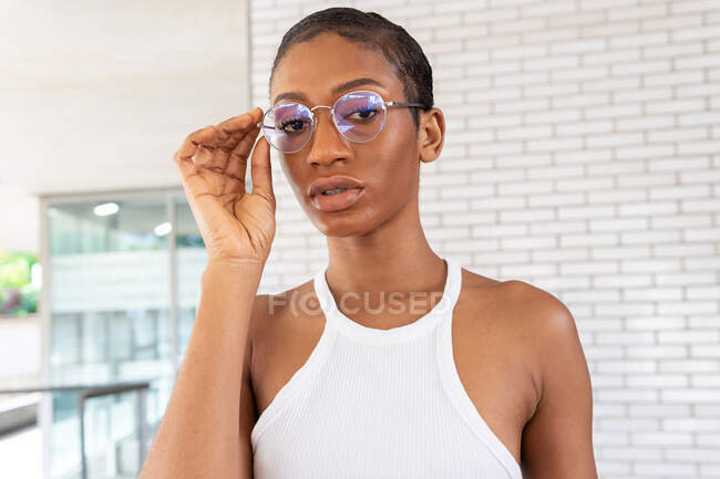 Fiduciosa donna afroamericana con i capelli corti in abito elegante con occhiali alla moda in piedi sulla strada vicino al muro di mattoni bianchi — Foto stock