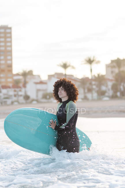 Vista lateral de la joven surfista en traje de neopreno en agua de mar ondulada con tabla disfrutando del día de verano - foto de stock