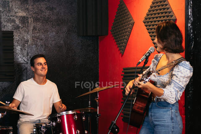 Positive young male playing drums near female musician playing acoustic guitar and singing while looking at each other in light club — Stock Photo