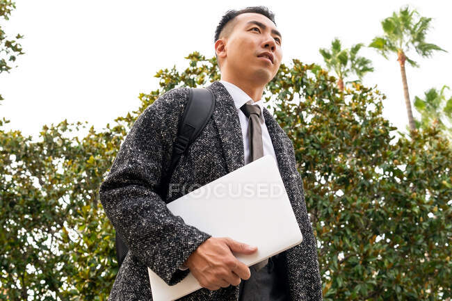 From below side view of contemplative young ethnic male entrepreneur with rucksack and netbook looking away against trees — Stock Photo
