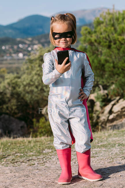 Menina auto-assegurada no traje de super-herói máscara olho com capa de navegação no celular — Fotografia de Stock