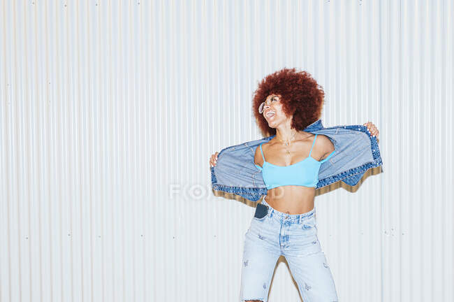 Joyeux femme mince avec coiffure afro portant des vêtements en denim élégant avec dessus et lunettes de soleil debout sur fond blanc sur la rue — Photo de stock