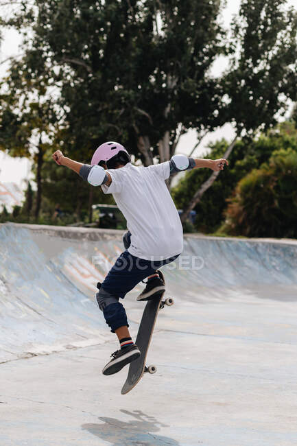 Все тело молодого этнического человека в повседневной одежде в защитном шлеме с наколенниками и локтевыми подушками на скейтборде в скейт-парке — стоковое фото
