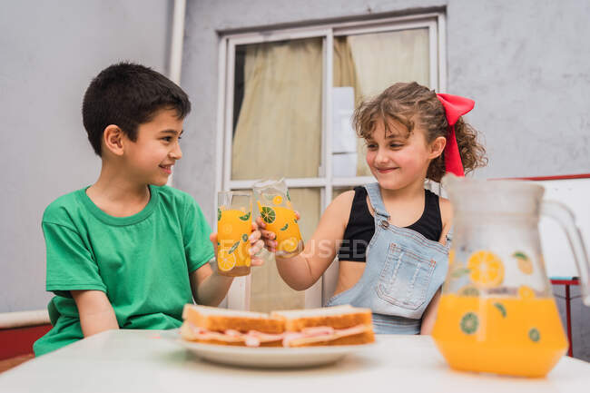 Crianças positivas com copos de suco doce sentadas à mesa com sanduíches frescos na placa na sala de luz em casa — Fotografia de Stock