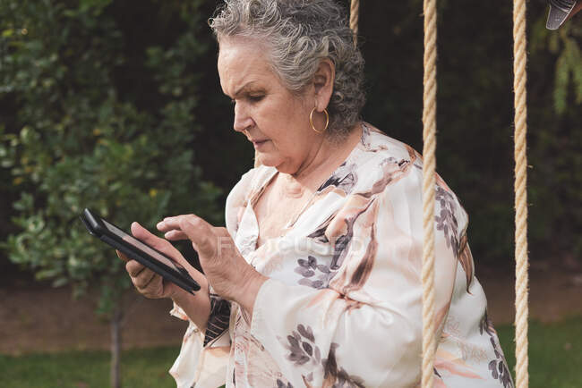 Seitenansicht einer Seniorin mit Bluse, die im Park sitzt und elektronisches Buch liest — Stockfoto