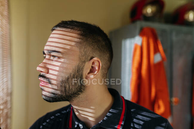 Crop side view of young bearded ethnic male wearing fireman sweat jacket looking out window and in bright sunlight — Stock Photo