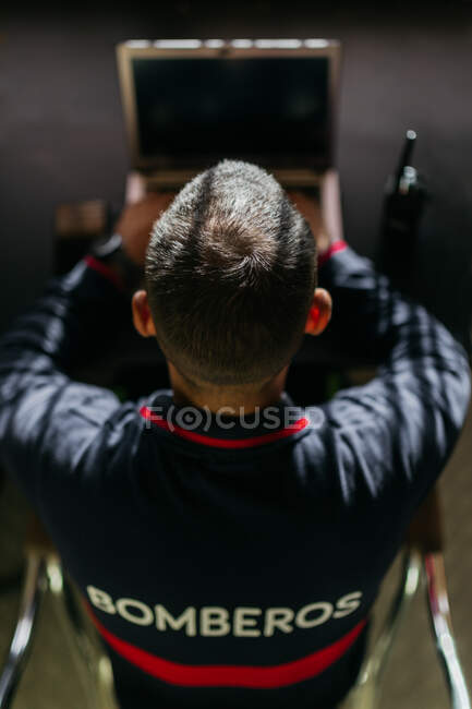 From above anonymous young ethnic firefighter with his back turned sitting at a table with a laptop and a transmitter looking at the screen — Stock Photo