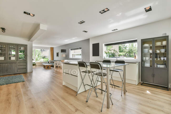 Stühle am Tisch im stilvollen Küchenbereich mit grauen Schränken mit verschiedenen Geschirr in modernen hellen Wohnung mit Fenstern platziert — Stockfoto