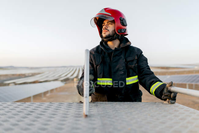 Firefighter wearing red protective helmet and uniform standing on fire truck ladder and looking away — Stock Photo