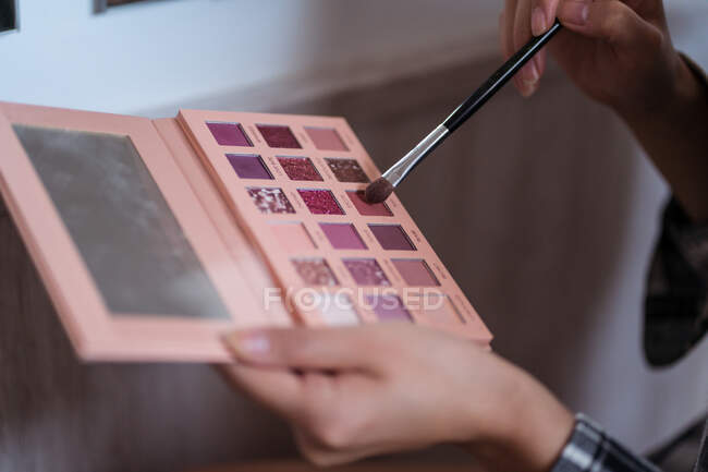 Crop anonymous female using cosmetic brush and eyeshadow palette with mirror in daylight on blurred background — Stock Photo