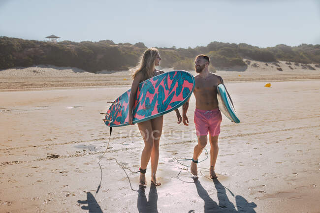 Full body of sportive couple in swimwear with surfboards looking at each other while standing on sandy seashore in tropical resort — Stock Photo