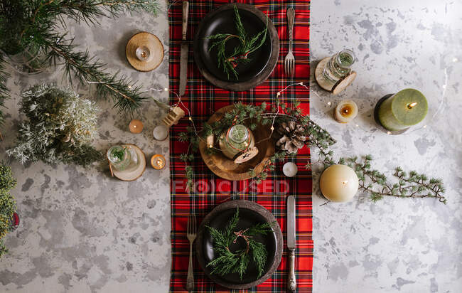 Vista superior de la mesa de Navidad con corona en el plato, adornos decorativos de madera y mantel a cuadros rojo con luces amarillas en el fondo - foto de stock