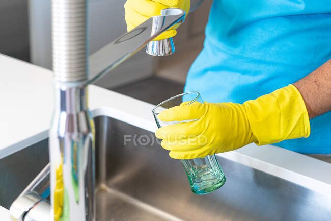 Анонимный мужчина в жёлтых резиновых перчатках наполняет стакан водой из-под крана во время уборки дома — стоковое фото