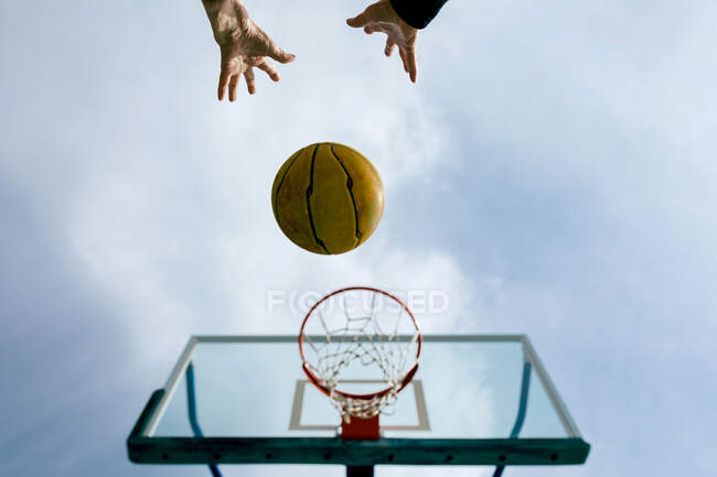 D'en bas de la culture personne anonyme jetant ballon de basket-ball dans le cerceau tout en jouant jeu sur le terrain de sport public contre le ciel bleu — Photo de stock