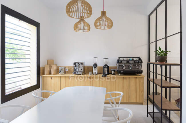 Macchine moderne per caffettiera e macinacaffè posizionate su bancone in legno in leggera caffetteria moderna con tavolino bianco vicino alla finestra e ripiani in legno — Foto stock