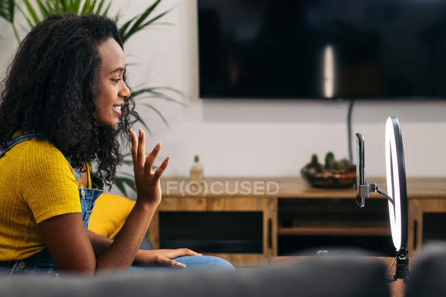 Sorridente femmina nera sul divano agitando mano durante l'utilizzo di smartphone sulla lampada ad anello a LED vicino a luci professionali su treppiedi — Foto stock