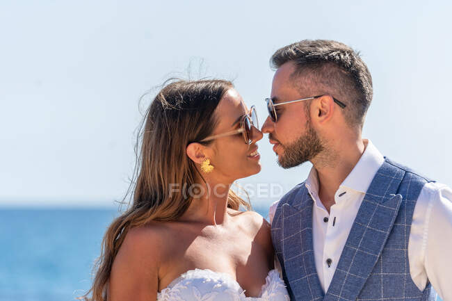 Вид сбоку веселая свадебная пара, стоящая на берегу у волнистого моря, наслаждаясь свадьбой в солнечной природе — стоковое фото
