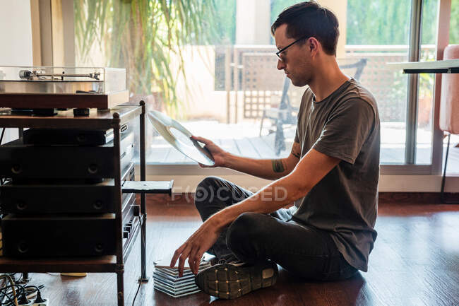 Вид сбоку мужчины в повседневной одежде, сидящего на полу рядом с поворотным столом и смотрящего на виниловую пластинку в руке — стоковое фото