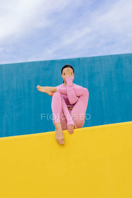 Corpo completo di donna in abbigliamento sportivo seduta con il viso chiuso a mano e una gamba piegata in posa Baby Cradle mentre pratica Hindolasana su sfondo blu e giallo — Foto stock