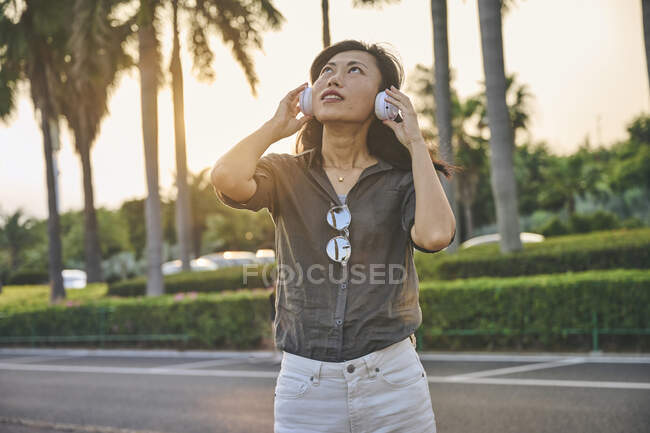 Seriöse Asiatin mit modernen weißen Kopfhörern blickt in die Ferne, während sie in der Nähe einer Straße in der Stadt mit grünen Bäumen steht — Stockfoto